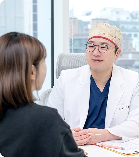 수술모를 쓴 흰 가운의 의사가 여성 환자와 마주 앉아 상담하는 모습
